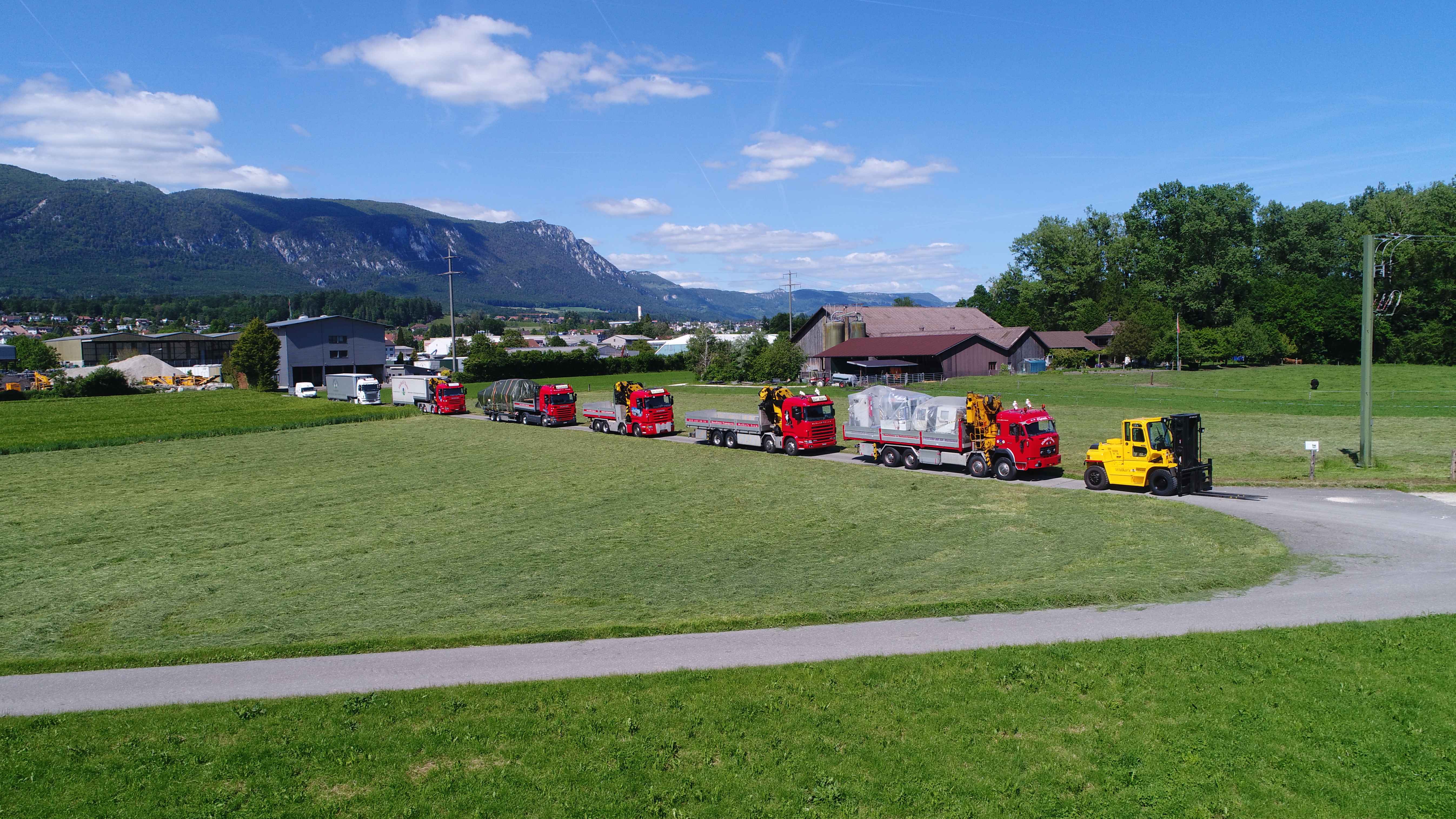Walker's Team fleet overview - multiple red trucks and yellow forklift lined up in scenic Swiss landscape with mountains and green fields